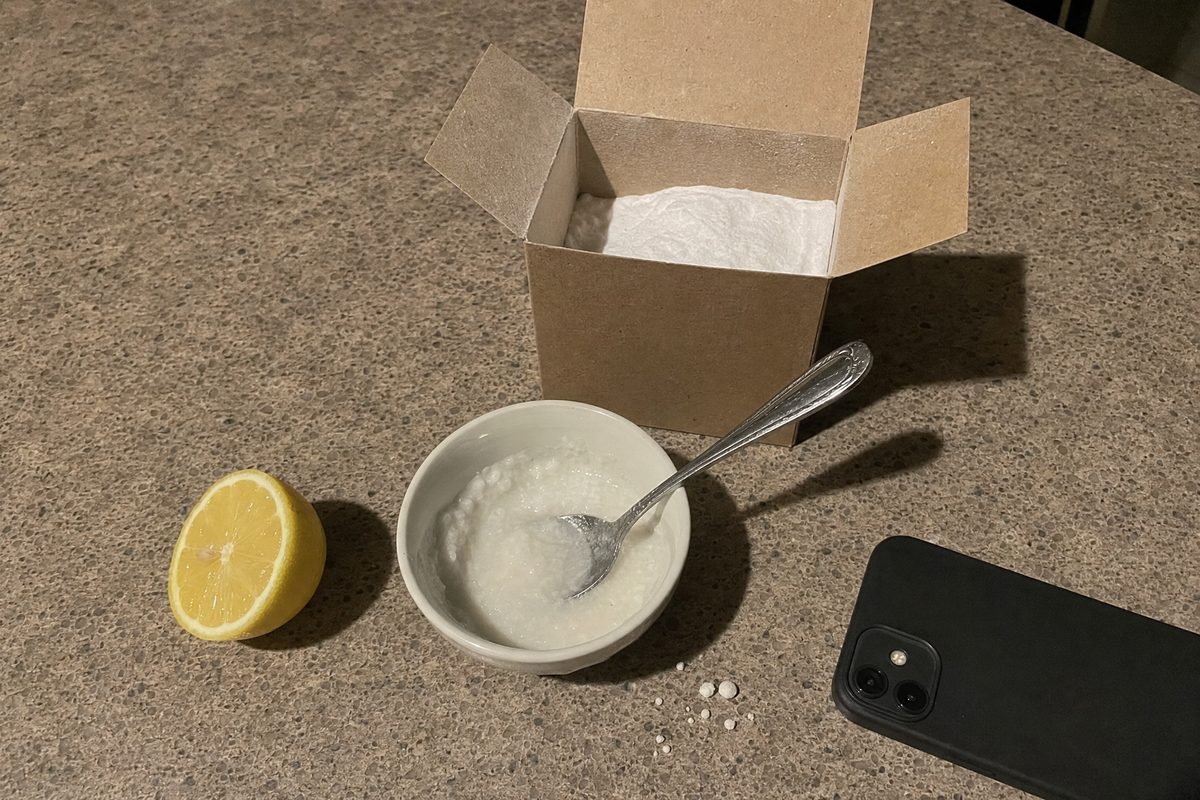 A small bowl of gritty white paste with a spoon, an open box of baking soda and a halved lemon on a kitchen counter