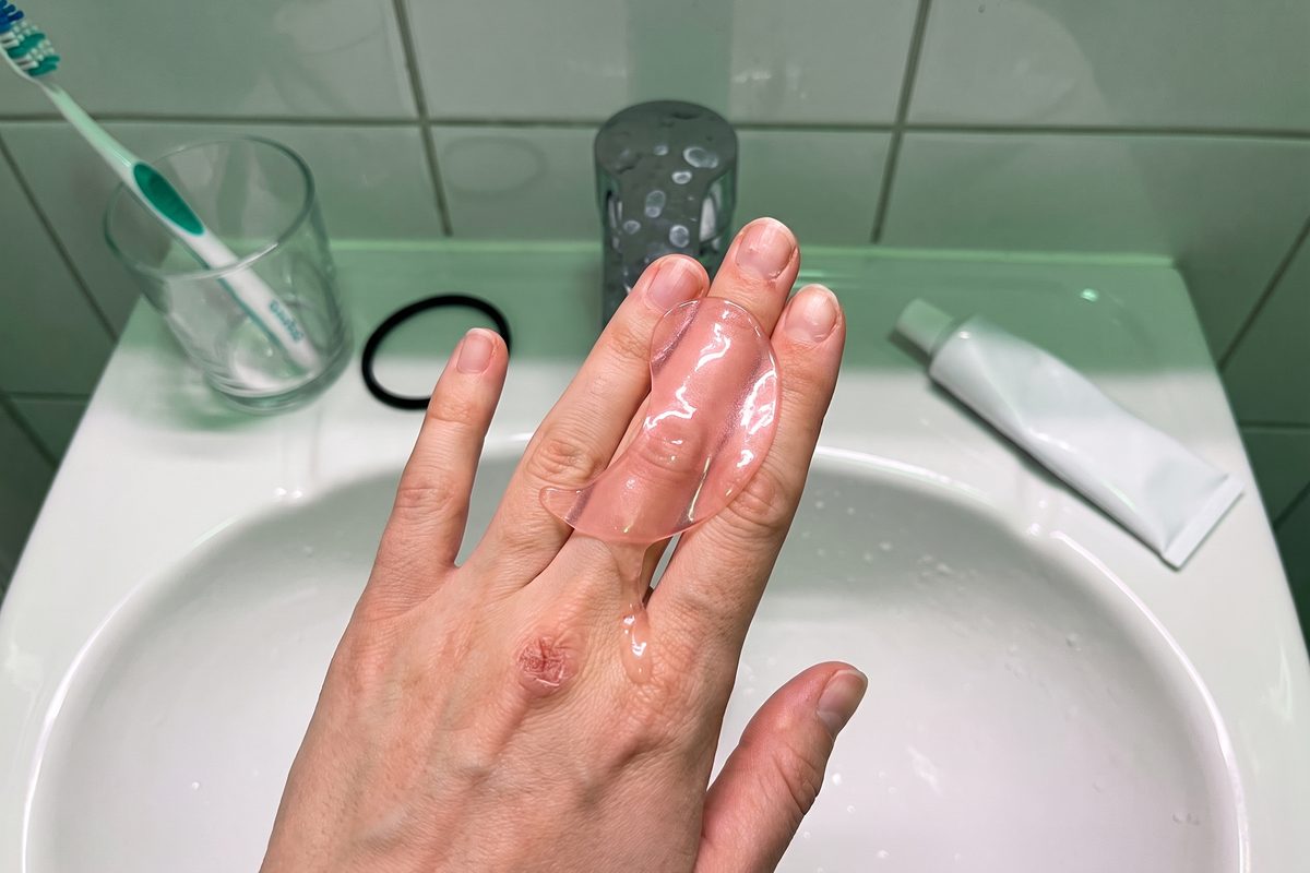 Close-up of a glossy pink half-moon hydrogel patch draped over a fingertip above a bathroom sink