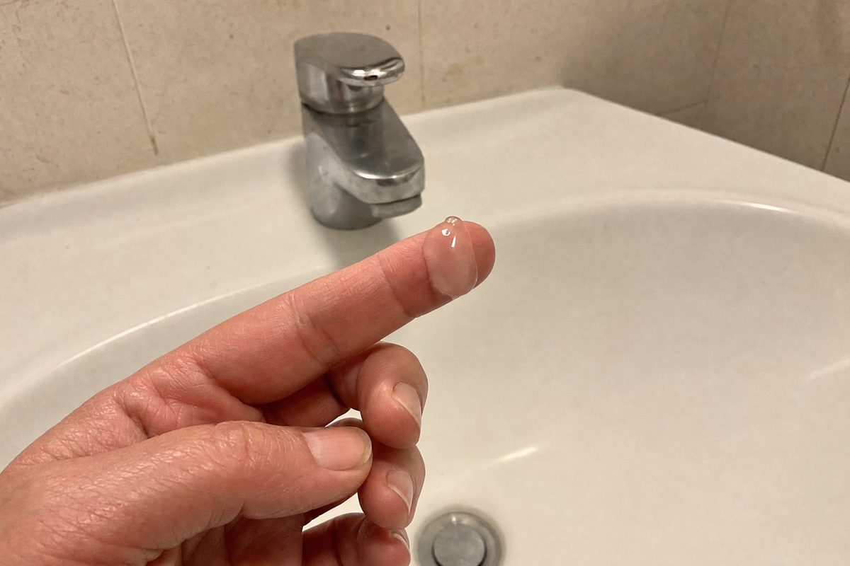 A glossy drop of clear gel on the fingertip of a woman's hand, early fifties, held above a bathroom sink