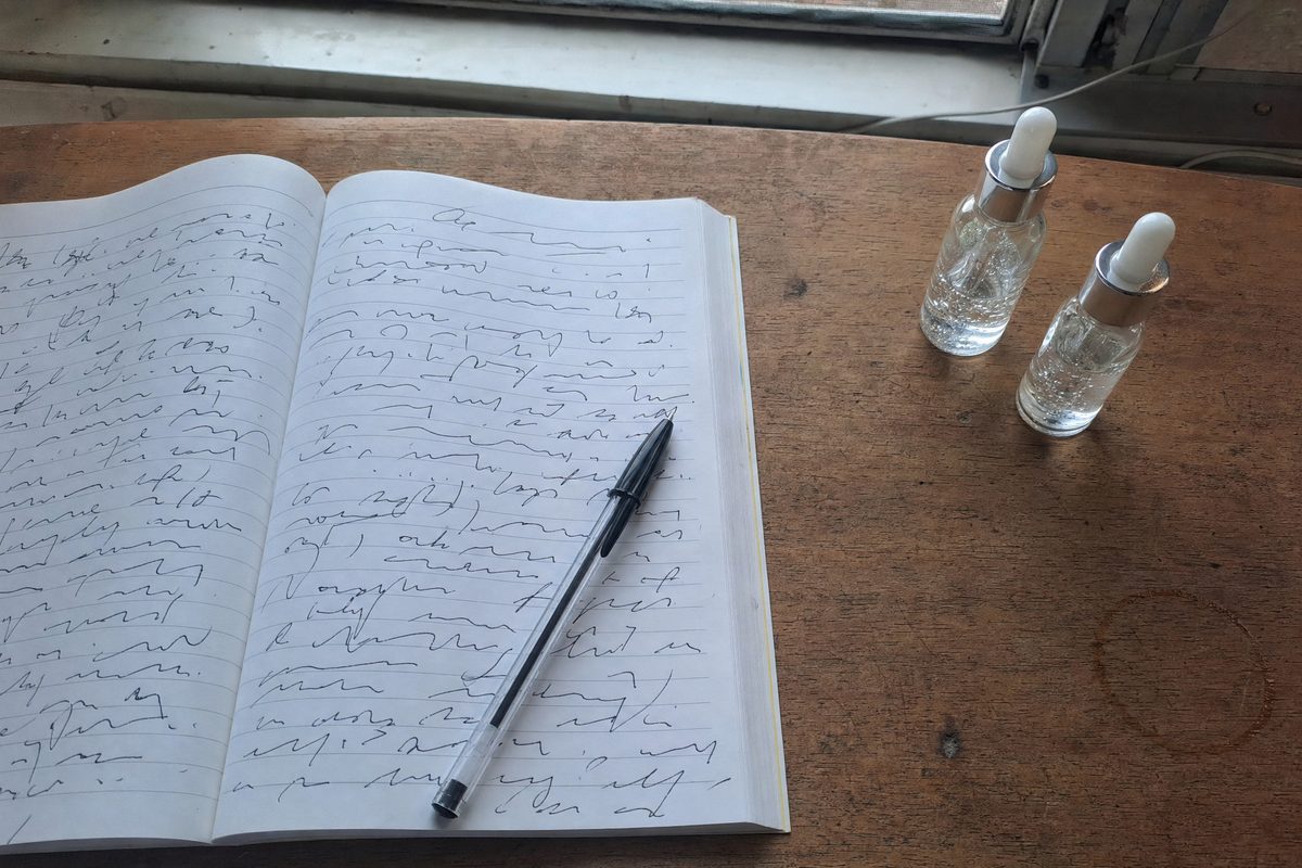 An open lined notebook with handwritten notes next to two small glass dropper bottles of clear gel on a wooden desk by a window