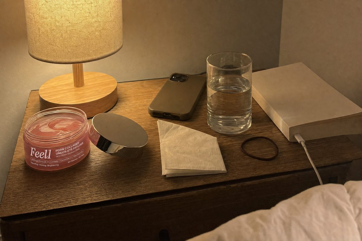 A frosted Feel1 PDRN eye patch jar with the lid off on a nightstand next to a phone and a folded tissue under a bedside lamp