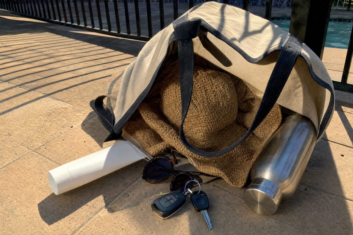A canvas beach bag tipped over on warm paving stones with a sunscreen tube, folded sun hat, sunglasses and a water bottle spilling out