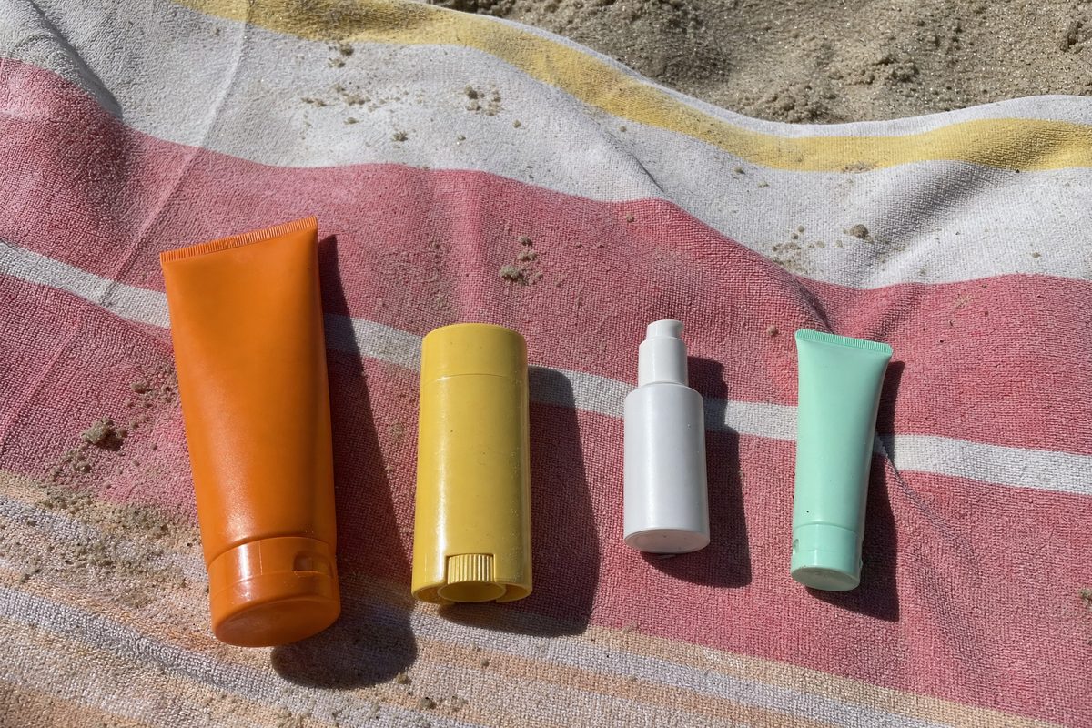 Four sunscreen tubes of different shapes and colours lying on a striped beach towel in hard midday sun, labels turned away