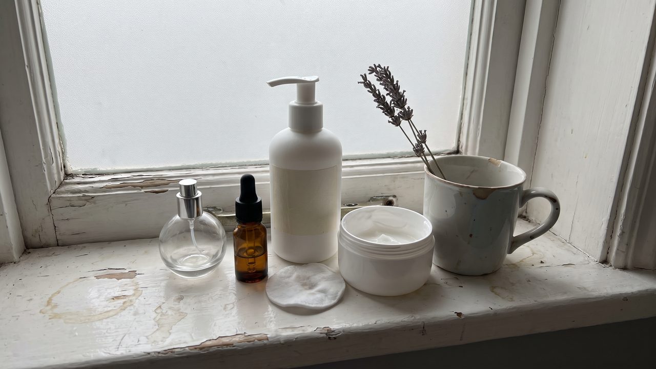 A bathroom windowsill with a perfume atomizer, a small amber essential-oil bottle, a pump bottle of lotion, an open tub of cream and a sprig of dried lavender