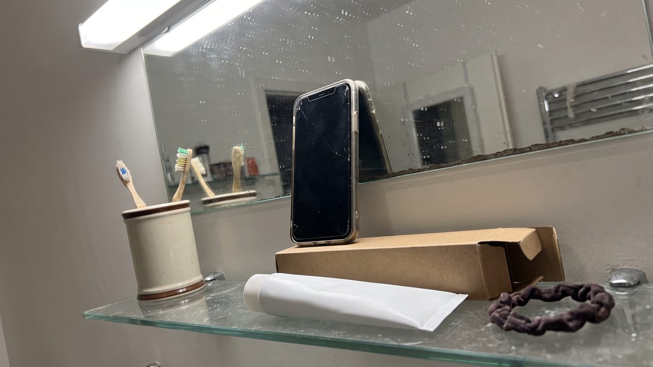 A bathroom mirror with a phone propped against it and a single new tube of cream on the shelf below