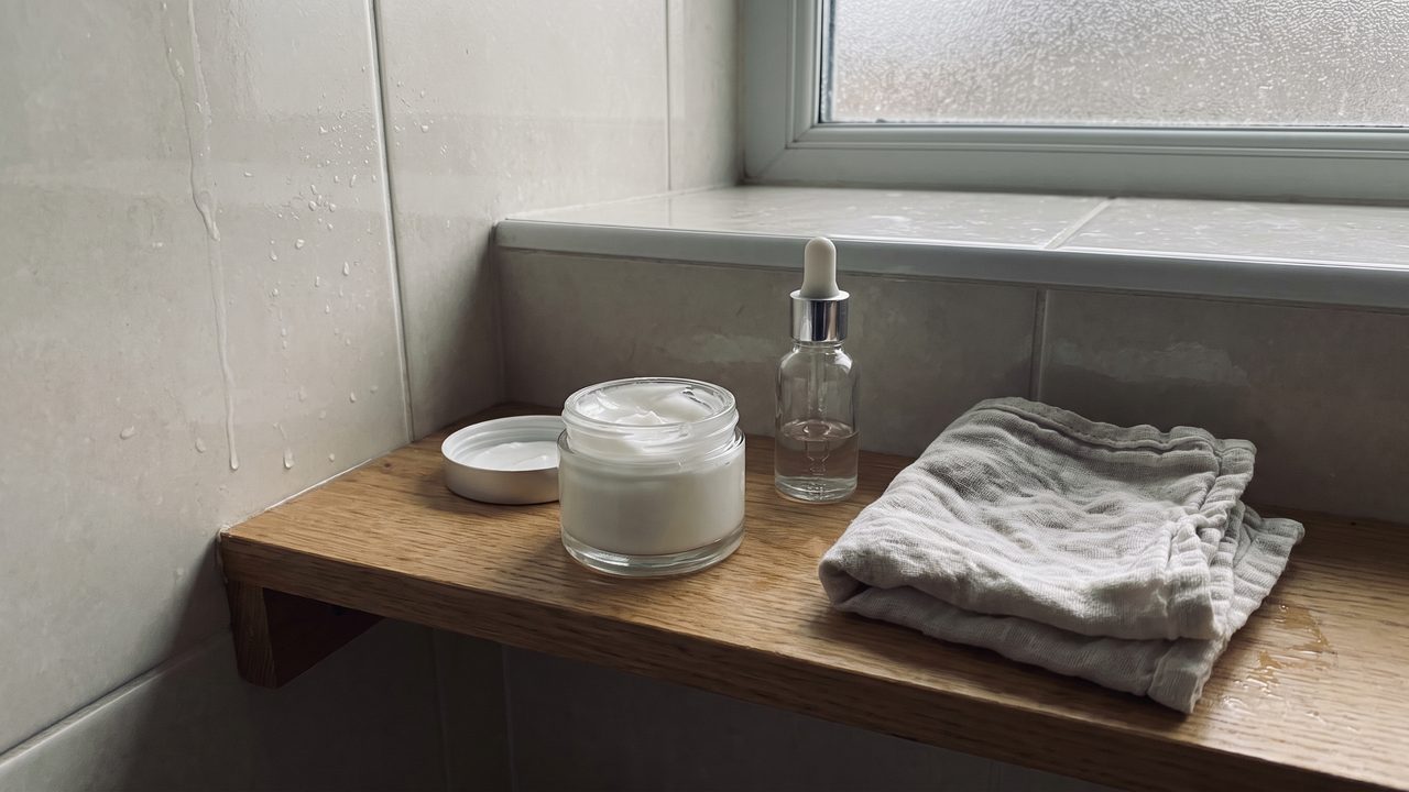 A serum bottle and an open jar of cream on a damp bathroom shelf