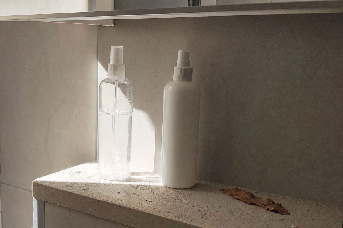 A clear spray bottle standing in direct sun on a bathroom shelf beside an opaque white bottle in the shade