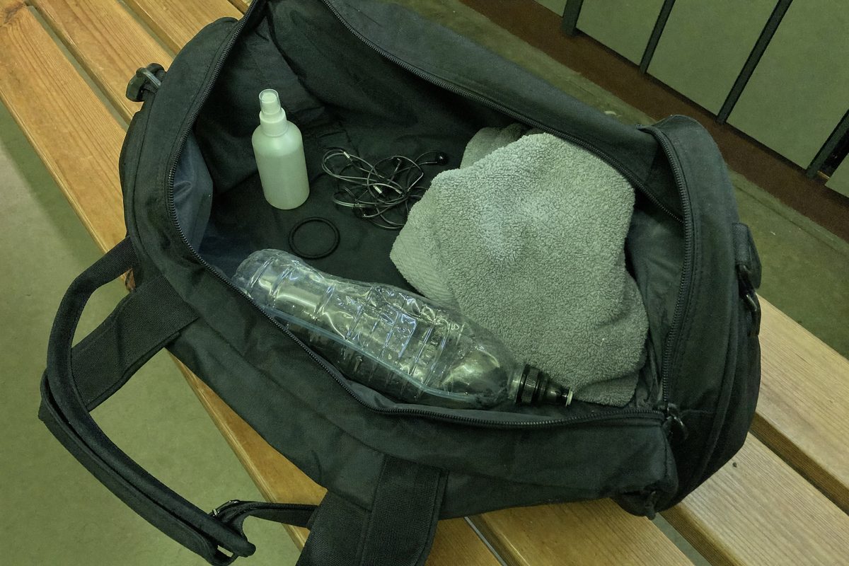 A small spray bottle, a rolled towel, earphones and a water bottle inside an open gym bag on a locker room bench