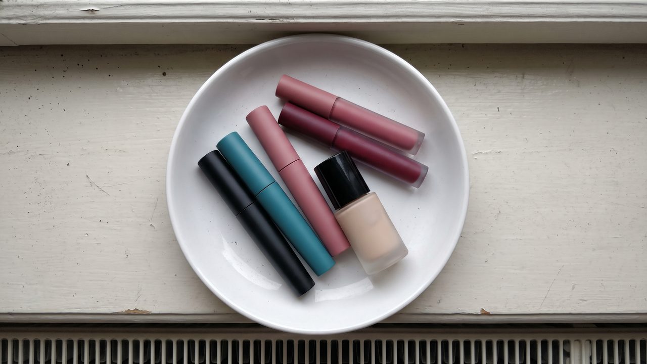 Six makeup products in plain solid-colour packaging lying in a shallow ceramic dish on a windowsill, photographed from above