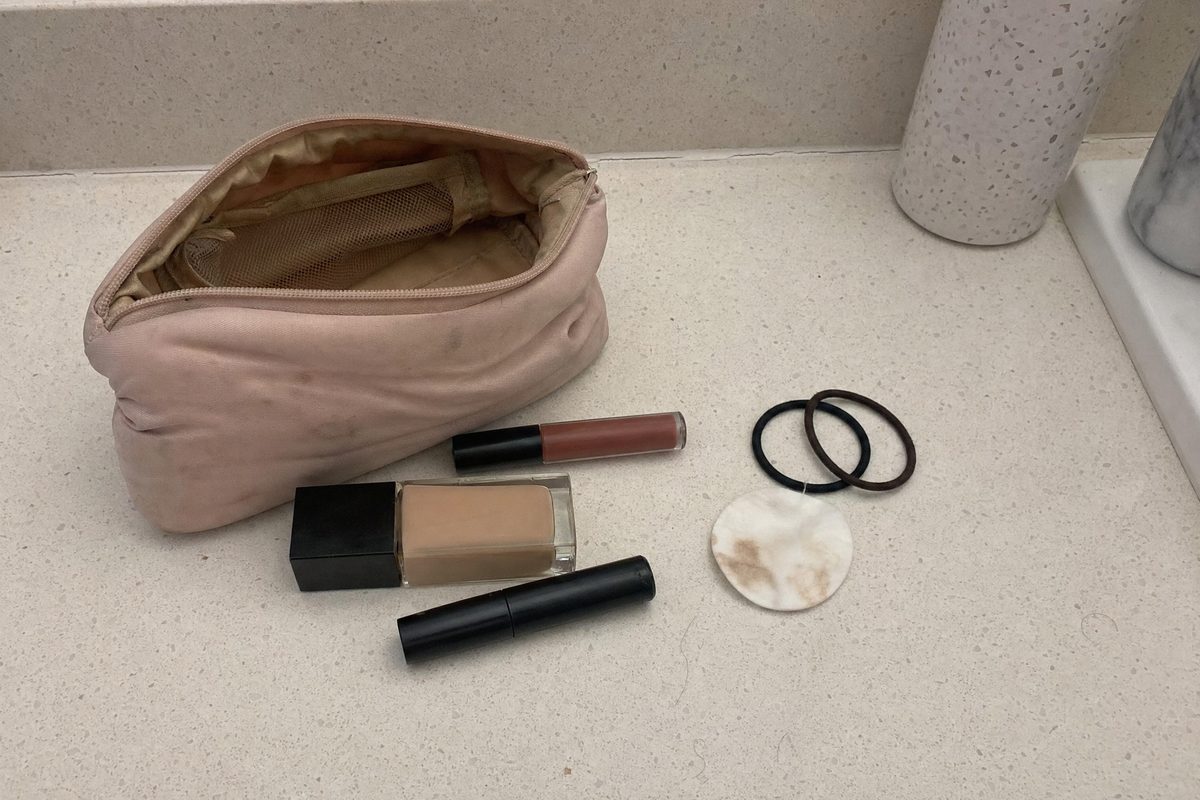 An open cosmetics bag spilling mascara, a liquid lipstick and a foundation bottle onto a bathroom counter under a ceiling light, labels turned away