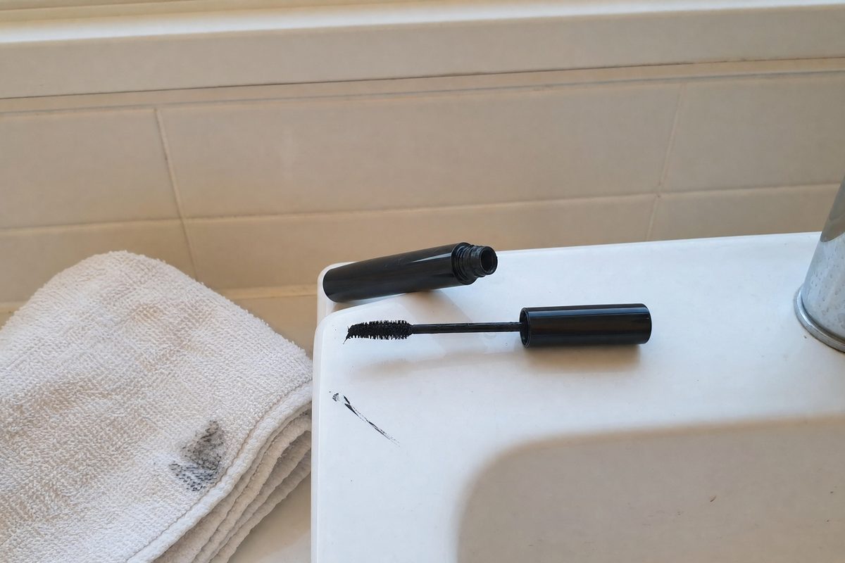 A mascara wand resting on the edge of a white bathroom sink next to a black smudge and a folded washcloth