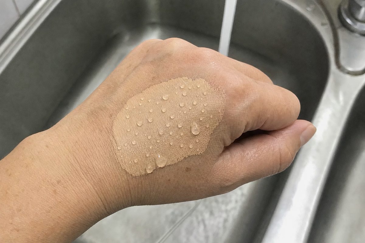 Water droplets beading on a swatch of liquid foundation on the back of a woman's hand, early fifties, held over a kitchen sink