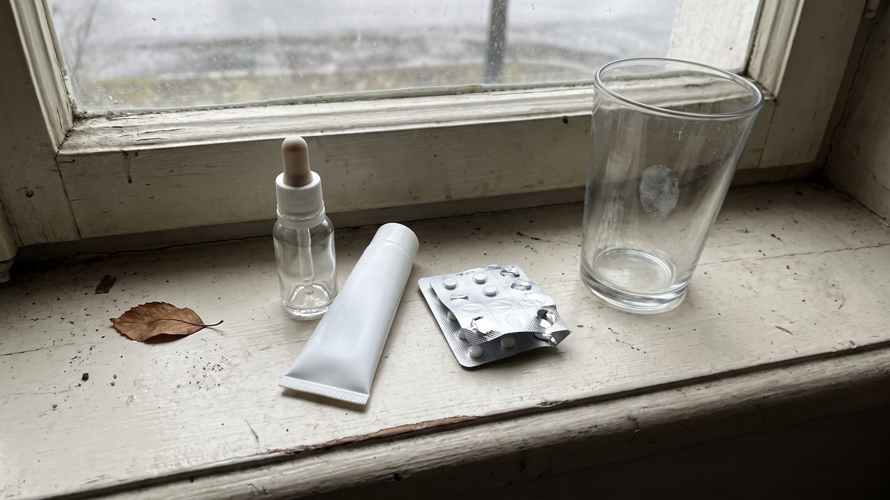 A small dropper bottle and a tube of cream on a windowsill next to a folded blister pack of tablets
